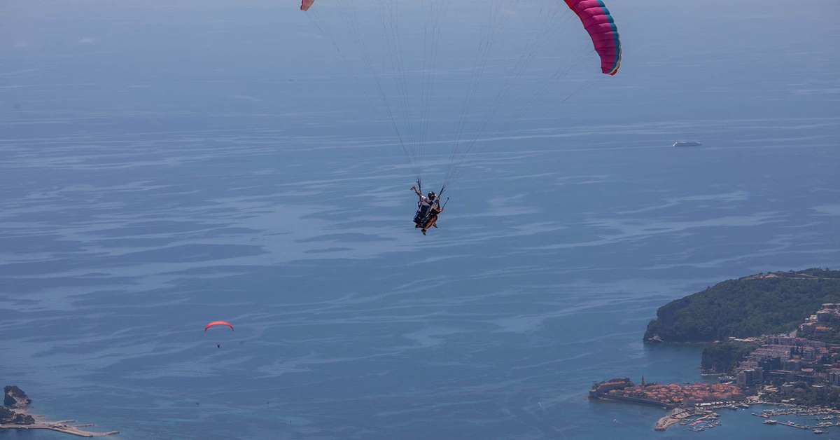 Tandem paraglider flying over the Adriatic Sea with Budva Old Town and the marina in the background.
