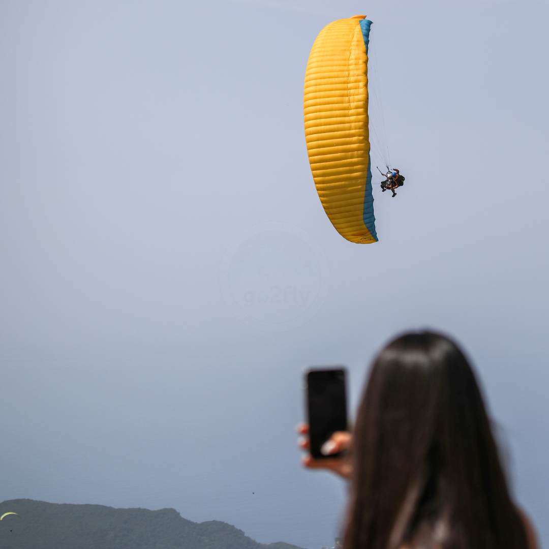 A tourist taking a photo of a yellow tandem paraglider performing a maneuver in Budva, Montenegro.