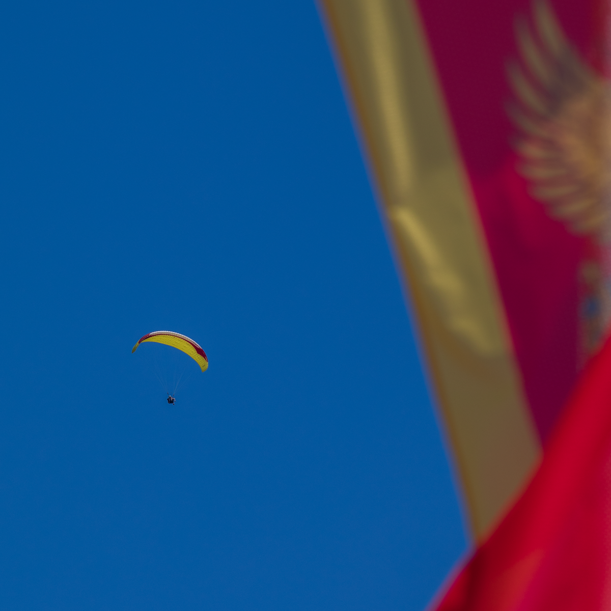 Paraglider with a yellow wing in a clear blue sky, partially obscured by the foreground of the red and gold Montenegrin flag.